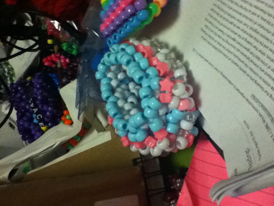 A pink, white and light blue kandi cuff made of pony beads on a cluttered desk, more beaded bracelets and papers around it